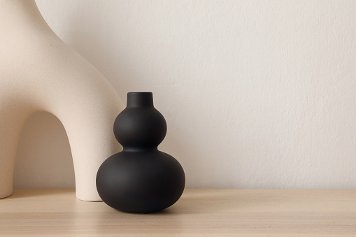 Set of modern ceramic vases of geometric organic shape on wooden table. Modern black sculpture. Beige wall. Abstract trendy artistic background, web banner.