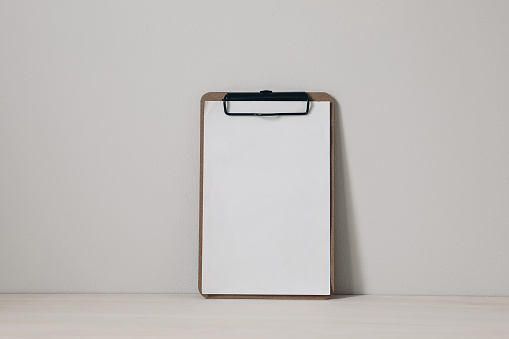 Wooden clipboard mockup with black clip and blank paper sheet on a wooden table against neutral beige wall background. Home office, creative desk. Elegant minimal interior, home decor, nobody.
