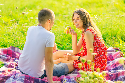 girl holds a pear and smiles at the guy. picnic in nature. couple on the meadow sits on a blanket.