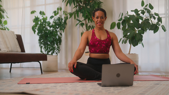 A Black ethnic young woman is using laptop and exercising in living room at home.