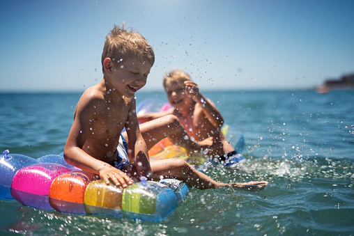 Two little boys are having fun on pool raft floating on the sea. Kids splashing and swimming fast.\nNikon D810