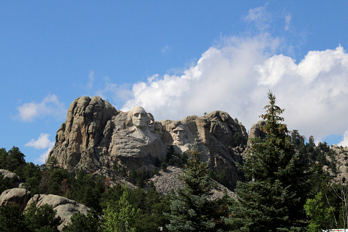 Color close-up photograph of the Mount Rushmore Monument with bright blue sky with big white fluffy clouds above and green trees in the foreground.