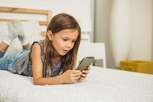 Little girl at her bedroom laying on bed and using smart phone. She is enjoying in her free time at home.