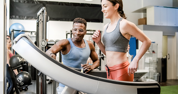 Side view of crop young fit lady in sportswear smiling while running on treadmill during workout with African American male personal trainer in sports club