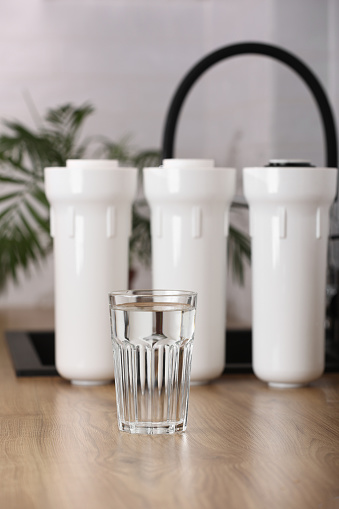 Glass of clean drinkable water and set of filter cartridges on wooden table top in a kitchen, houseplant.Installation of reverse osmosis water purification system. Concept household filtration system.