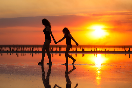 Happy family summer travel holiday. Silhouette of mom with child daughter holding hands walking together on beach on sunset. Happy mothers day. Concept of family values. International Children's Day.