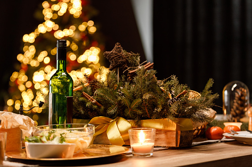 Close up shot of rustic set up on a dining table for a Christmas dinner party. Tea candles, beautiful center piece and salads served next to a bottle of wine. Christmas tree glistening in the background.