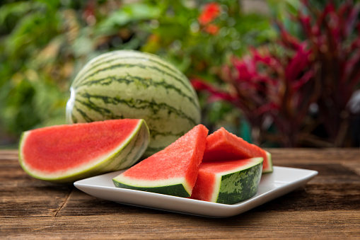 This is an outdoor photograph of sliced watermelon on a white square modern plate sitting on a wooden picnic bench outdoors for a simple and concept of healthy eating and snacks during the summertime fun.