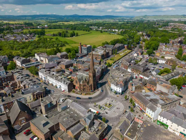 This aerial drone photo shows an old church in the city center of Dumfries, Scotland. There is a large square in front and some greenery in the background.