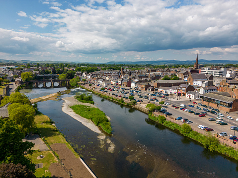 This aerial drone photo shows the city of Dumfries in Scotland, United Kingdom. There is a large river running through the town. The city center can be seen in the background.