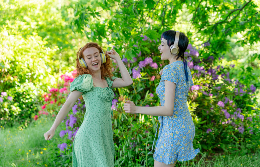 Outdoor portrait of two happy  women, girlfriends, friends in dresses and headphones dancing in the public park. Listen to online music anywhere and be yourself concept