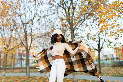 Young adult Black female in warm poncho relaxing in autumn park