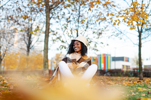 Young adult Black female in warm poncho relaxing in autumn park