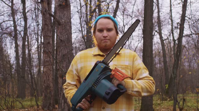 A red lumberjack picks up a chainsaw in the autumn forest and looks into the camera