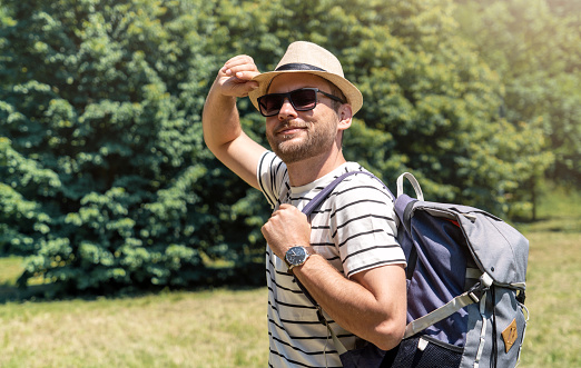 Adult man with sunglasses, hat, and backpack enjoying summer in the city park, looking at camera and smiling.