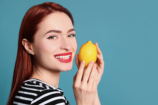 Happy woman with red dyed hair holding whole lemon on light blue background, space for text