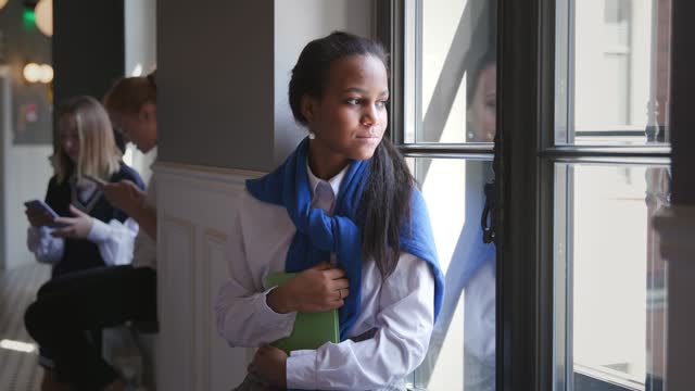 Portrait of sad African-American teen student sitting on window sill in corridor.