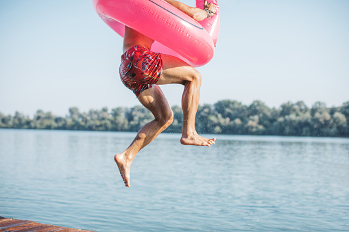 Young man jumping in river from deck. He is jumping with pink flamingo swimming float.