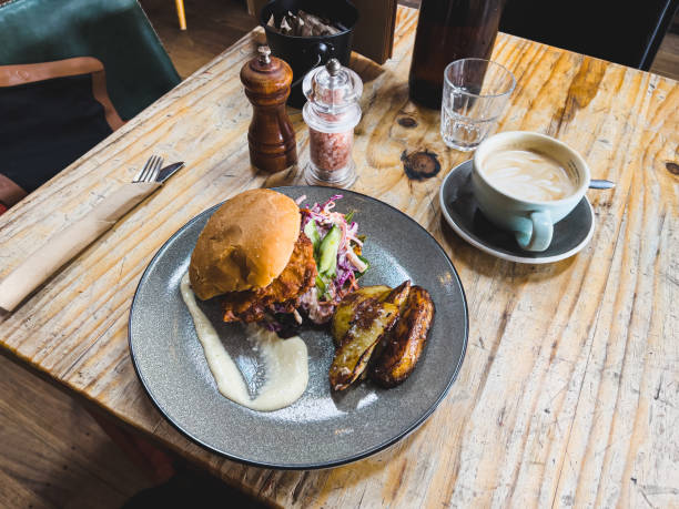 Horizontal high angle closeup photo of a Southern Fried Chicken Burger with red cabbage coleslaw, tomato chutney, mayonnaise and roasted potato wedges, served on a grey ceramic plate on a rustic wooden table in a restaurant for lunch. Salt and pepper grinders, cutlery, paper serviette and a cup of latte coffee on the side. Uralla near Armidale, New England high country, NSW.