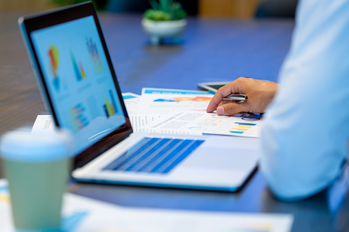 Close up of Business man working with a laptop and documents with financial charts graphs and finance figures. He is holding a pen. He is in an office