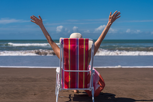 Woman raising her hands up while relaxing sitting on a deck chair at the beach. Summer concept.