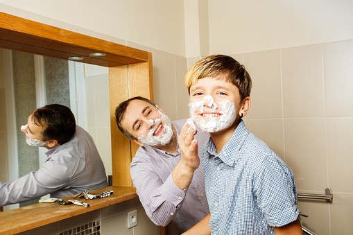 Dad and kid preparing to shave together foaming face smiling at camera in bathroom