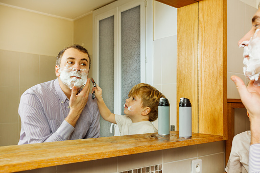 Father shaving looking at mirror in bathroom with kid holding razor helping to shave