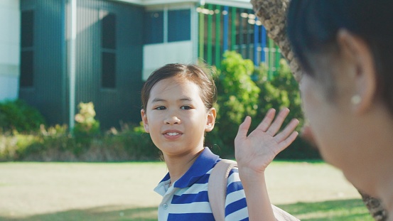 Mixed Asian primary school student girl waving goodbye to her Asian grandmother at school on sunny day, active travel, healthy lifestyle