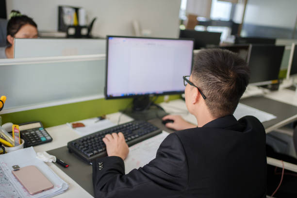 A business professional in a suit is seen writing notes on a clipboard while using a computer. The setting reflects a focused and organized workspace.