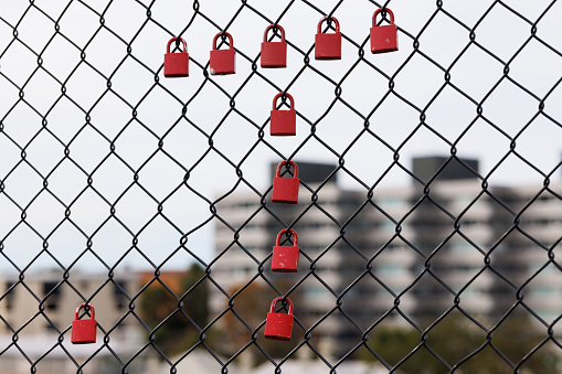Red metal padlocks attached to a fence with buildings beyond.