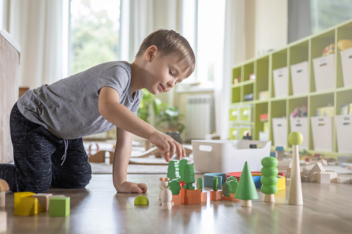 Confident male kid building fortress tower architecture wooden bricks ecology Montessori material. Baby boy playing construction eco friendly rainbow bricks at nursery childish room home toys storage