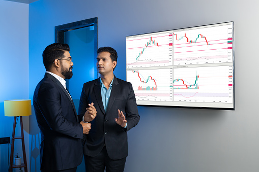 Indian businessmen colleagues trading and discussing stocks online in office. Stock brokers looking at indexes graphs on big computer screen. Crypto and forex trading.
