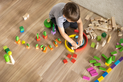 Confident male kid building fortress tower architecture wooden bricks ecology Montessori material. Baby boy playing construction eco friendly rainbow bricks at nursery childish room home toys storage