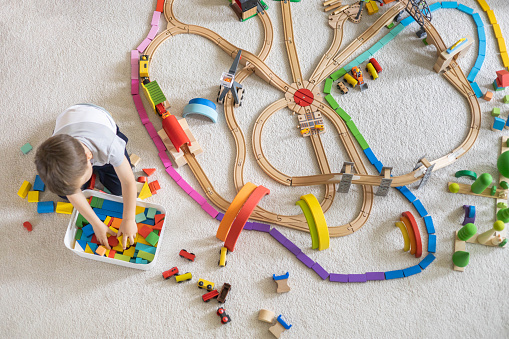 Preschool baby boy playing wooden Montessori materials rainbow arch railways at childish room. Playthings storage cupboard arrangement organizing. Male kid with natural eco friendly toys at home. Top view.