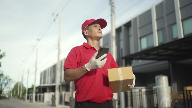A parcel delivery worker wearing a red uniform is holding a parcel box to the recipient. At that time he was using a mobile phone. contact the receiver in front of the house