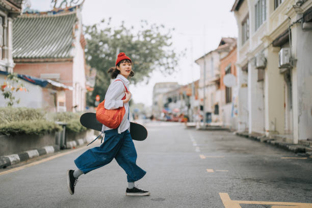 szczęśliwa azjatycka chinka przechodząca przez ulicę niosąca deskorolkę na starym mieście - teenager youth culture city life fashion zdjęcia i obrazy z banku zdjęć