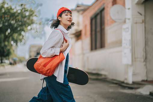 Happy Asian Chinese young woman crossing road carrying skateboard in old town