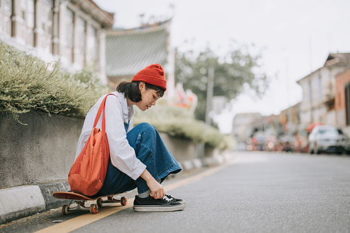 Asian Chinese young woman tying shoelace at roadside sitting on skateboard