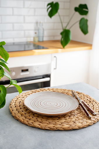 Wooden blue table top with flower pot and plate for breakfast against blurred white kitchen with cutting board in scandinavian style in morning light. Vertical orientation.