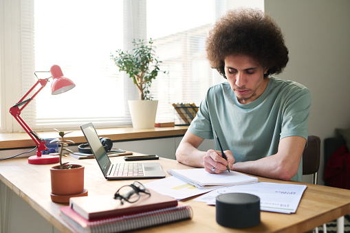 Young man sitting at table with gadgets and making notes in notepad, he studying at home