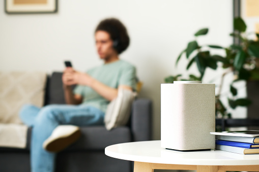 Close-up of smart speaker standing on coffee table in the room with man using smartphone in background