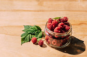 Red raspberry in a jar, wooden background, summer seasonal food, bright sunlight and shadows. Background of fruity ripe berries