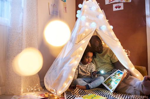 Dad reading book to his child while they sitting in handmade tent in the room