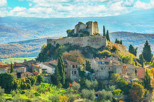 The photo depicts the picturesque Italian village of Castiglione d'Orcia, located in the region of Tuscany, Italy. The village is surrounded by beautiful rolling hills and countryside. Sitting prominently on top of a hill is a derelict ruin (Rocca Aldobrandesca), which adds a touch of mystery and history to the scene. The sunlight bathes the village in a warm glow, illuminating the rustic buildings. The blue sky overhead complements the earthy tones of the village, creating a serene and timeless atmosphere.