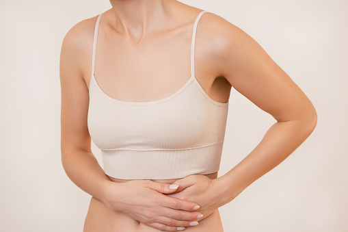 a young woman in beige underwear holds her hands by the side of her stomach, experiences sharp severe pain, isolated on a beige background. The concept of health