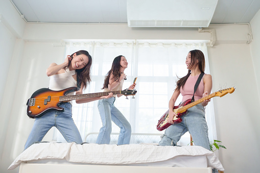 Group of Beautiful Asian women playing electric guitar and electric bass and singing on bed. Cheerful Pretty Girls rock musician enjoying the music in bedroom. Females friends having fun.