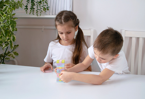 Two children are playing with colorful cubes at white table. Brother and sister are playing board games at home and pulling wooden cubes out of the tower. Concept of children's leisure at home.