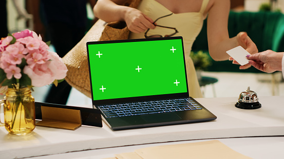 Laptop with greenscreen template on hotel reception counter next to lounge area, isolated display mockup template at front desk. Blank chromakey copyspace on pc in resort lobby.