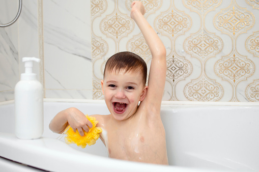 Funny child is having fun bathing, sitting in white bath with soap and shampoo foam and washing his head with his hair. Shampoo, hair care product and soap for children. Concept of health and hygiene.