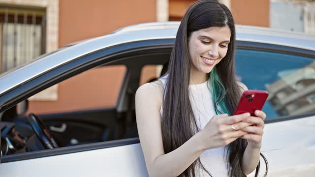Young beautiful hispanic woman using smartphone leaning on car at street